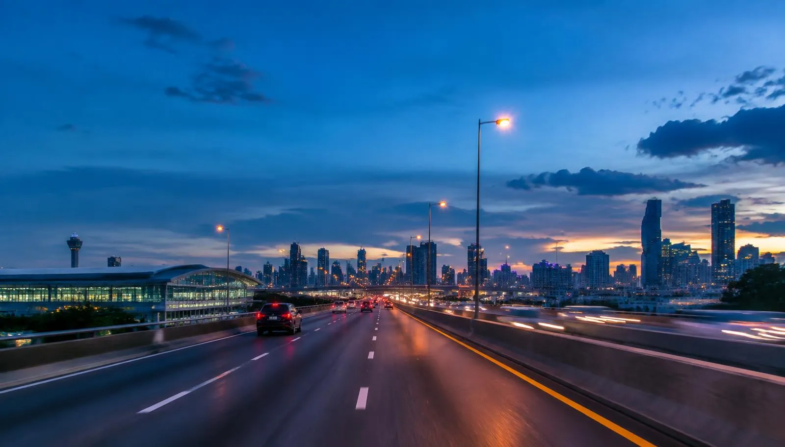 Elevated expressway at dusk with light traffic heading toward a city skyline, an airport terminal on the left